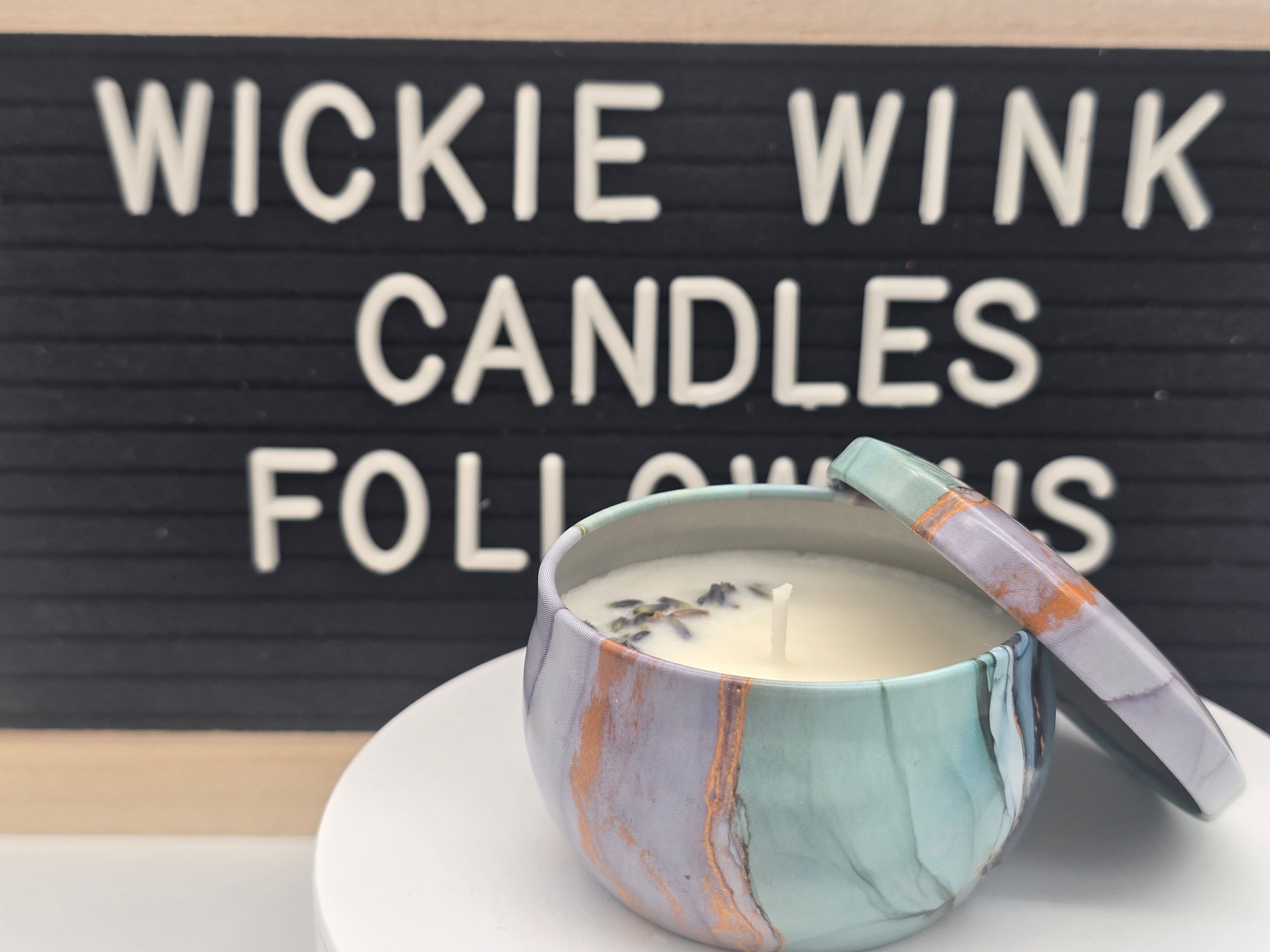 Candle in a decorative container with 'Wickie Wink Candles' sign in the background