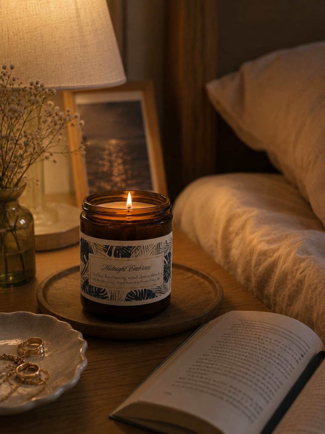 Candle with a label on a wooden table next to an open book and a lamp in a cozy room.